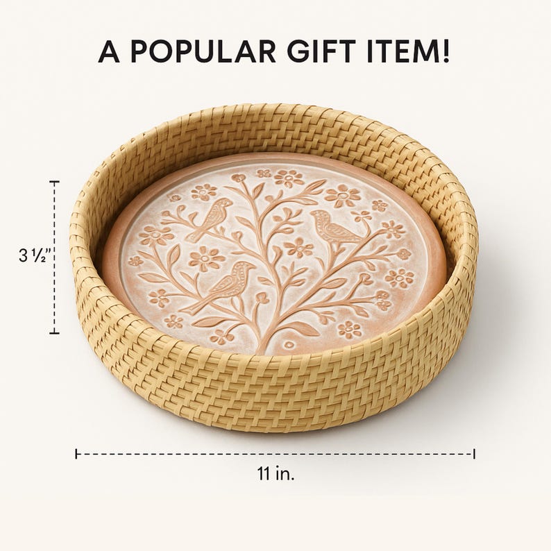 Op de afbeelding: Ronde decoratieve schaal in terracotta kleur met een vogel- en bloemmotief, geplaatst in een geweven mand. De schaal heeft een diameter van 28 cm en is 9 cm hoog. De tekst "A POPULAR GIFT ITEM!" staat bovenaan.