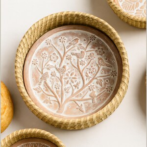Op de afbeelding: Een ronde, terracotta-kleurige decoratieve schaal met een geweven, lichtbruine rand. De schaal heeft een reli&euml;f ontwerp van een boom met vogels en bloemen. De schaal is omgeven door een geweven, lichtbruine rand.