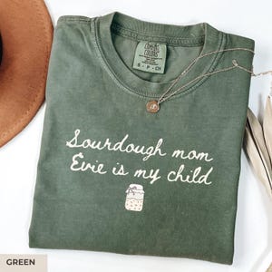 May include: Green t-shirt with the text "Sourdough mom Evie is my child" in white script. A small jar illustration is below the text. A silver necklace with a pendant is on the shirt. The shirt is labeled "Comfort Colors" and is size S-CH.