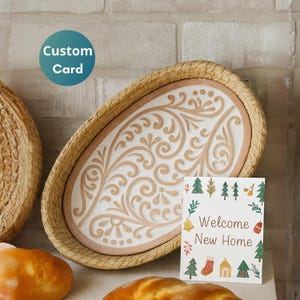 May include: An oval decorative tray with a woven border and a white interior featuring a raised, swirling design in a light brown color. A greeting card with the text "Welcome New Home" is displayed in front of the tray. A blue circle with the text "Custom Card" is in the upper left.