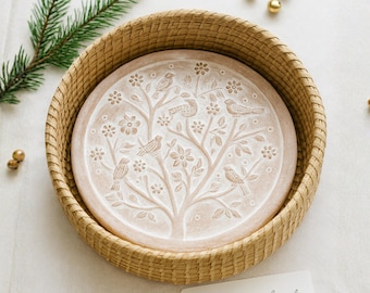 May include: A round, beige ceramic bread warmer with a tree and bird design, nestled in a woven basket. The warmer features a detailed relief of a tree with birds and flowers. The basket is light brown. A sprig of greenery and gold ornaments are in the background. The text on the card reads: "Where warm bread meets home."