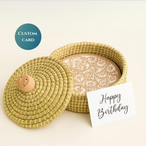 May include: A woven basket with a lid and a decorative floral design inside. The lid has a wooden knob. A card with "Happy Birthday" is next to the basket. A teal circle with "CUSTOM CARD" is in the upper left.