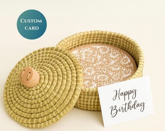 May include: A woven basket with a lid and a decorative floral design inside. The lid has a wooden knob. A card with "Happy Birthday" is next to the basket. A teal circle with "CUSTOM CARD" is in the upper left.