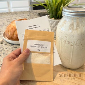 May include: A sourdough starter kit with a jar of active starter, a packet of dehydrated starter, and instructions. A loaf of bread sits on a plate in the background. The packet reads "Dehydrated Sourdough Starter 10g."