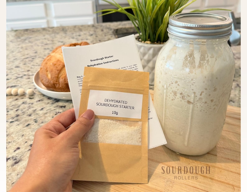 May include: A sourdough starter kit with a jar of active starter, a packet of dehydrated starter, and instructions. A loaf of bread sits on a plate in the background. The packet reads "Dehydrated Sourdough Starter 10g."