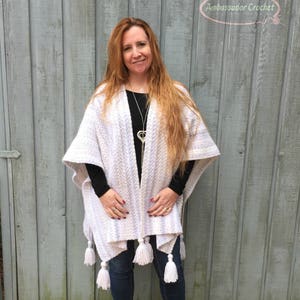 May include: A white crocheted poncho with white tassels. The poncho has a striped pattern with light purple stripes. The model is wearing the poncho with a black shirt and blue jeans.