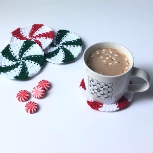 May include: A white mug with a black floral design sits on a red and white crocheted coaster. The mug is filled with hot chocolate topped with mini marshmallows. Four red and white peppermint candies are scattered on a white surface. Three green, white, and red crocheted coasters are in the background.