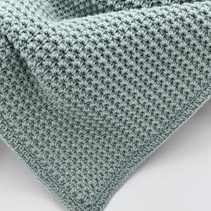 May include: A close-up of a handmade, light teal crochet blanket. The blanket features a textured stitch pattern, with a dense, woven appearance. The corner of the blanket is visible, showcasing the detailed craftsmanship and soft color.