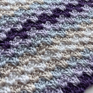 May include: A close-up of a crocheted blanket with stripes of purple, light blue, white, and beige yarn.