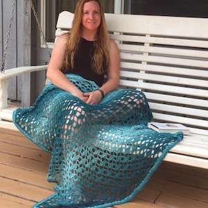 Puede incluir: Una mujer se sienta en un columpio de porche blanco con una manta de crochet turquesa drapeada sobre sus piernas. La manta tiene un degradado de color de turquesa claro a turquesa oscuro.