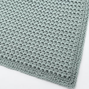May include: A close-up of a crocheted blanket in a soft, muted green color. The blanket features a textured stitch pattern, with a detailed, close-up view of the yarn's texture and the intricate design. The edges are neatly finished.