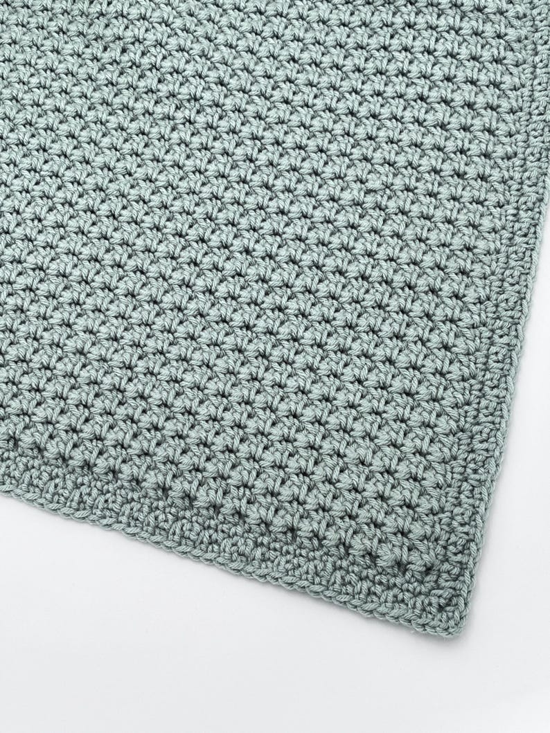 May include: A close-up of a crocheted blanket in a soft, muted green color. The blanket features a textured stitch pattern, with a detailed, close-up view of the yarn's texture and the intricate design. The edges are neatly finished.