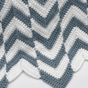 May include: A handmade crochet blanket with a chevron pattern. The blanket features alternating stripes of white and a muted blue-gray color. The edges of the blanket have a scalloped finish, adding a decorative touch to the overall design.