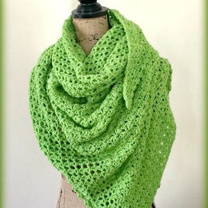 May include: A bright green, crocheted shawl draped over a mannequin. The shawl has a textured, open weave design and a scalloped edge. The text "Ambassador Crochet" is visible in the upper right corner.