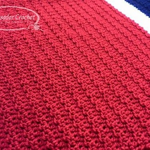 Puede incluir: Primer plano de una manta roja tejida a ganchillo con un patrón de puntadas texturizadas. La manta es rectangular y parece estar hecha a mano. Las palabras "Ambassador Crochet" están en la esquina superior izquierda.