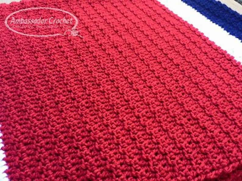 Pu&ograve; includere: Primo piano di una coperta rossa all'uncinetto con un motivo a punto strutturato. La coperta &egrave; rettangolare e sembra fatta a mano. Le parole "Ambassador Crochet" sono nell'angolo in alto a sinistra.