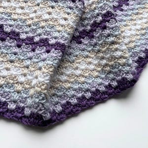 May include: A crocheted baby blanket in shades of purple, blue, white, and beige. The blanket is folded in half and has a textured stitch pattern.