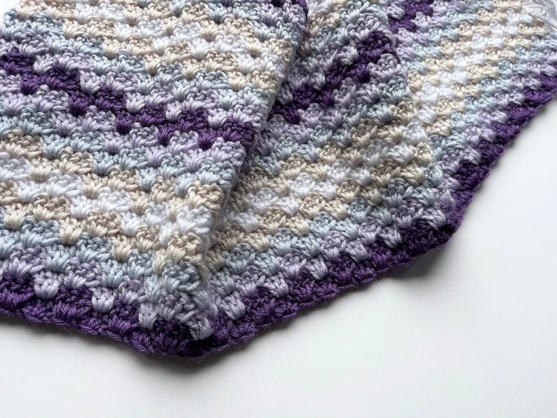 May include: A crocheted baby blanket in shades of purple, blue, white, and beige. The blanket is folded in half and has a textured stitch pattern.