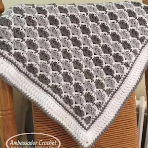 May include: A gray and white crocheted baby blanket with a diamond pattern. The blanket is folded and draped over a wooden chair.