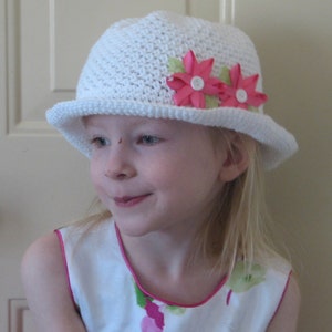 May include: A young girl wearing a white crocheted hat with pink flowers and a white dress with pink trim.