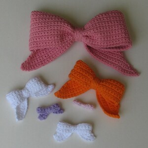 May include: Five crocheted bows in various sizes and colors. The largest bow is pink, the next largest is orange, and the three smallest are white, purple, and pink.