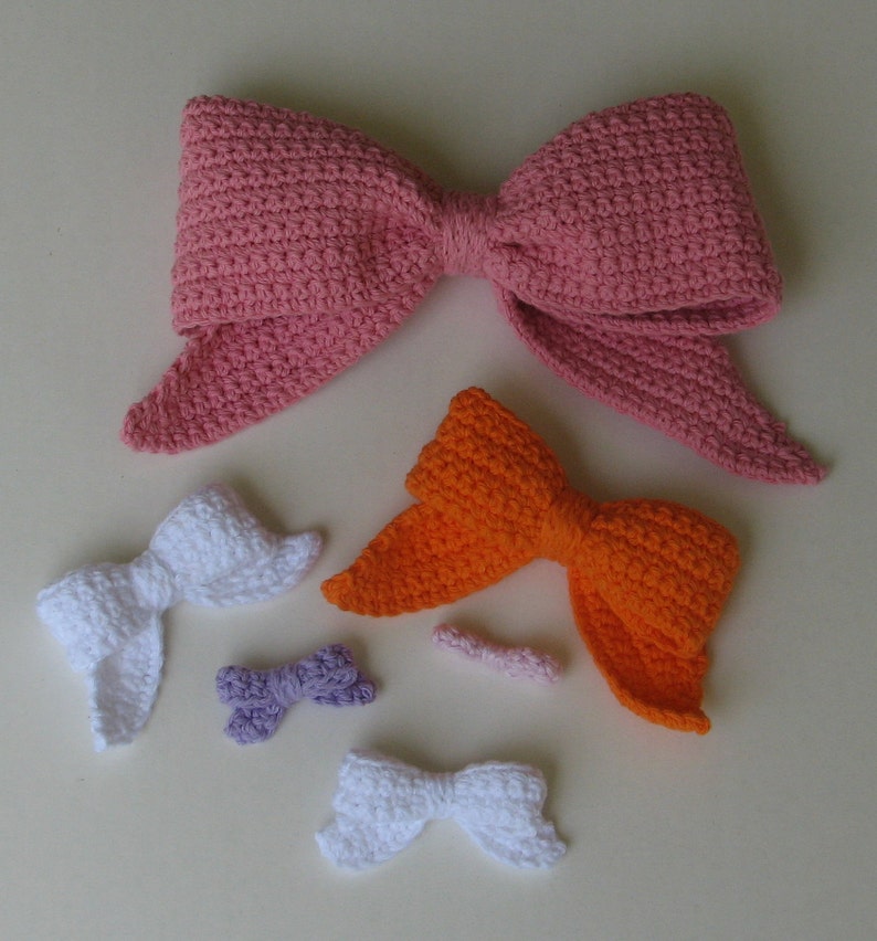 May include: Five crocheted bows in various sizes and colors. The largest bow is pink, the next largest is orange, and the three smallest are white, purple, and pink.