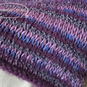 Puede incluir: Primer plano de una tela de crochet de color morado, azul y gris con una textura peluda. La tela está hecha con una variedad de puntadas y colores, creando un patrón único e interesante.