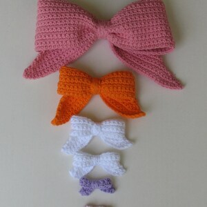 May include: A set of six crocheted bows in various sizes and colors, including pink, orange, white, purple, and light pink. The bows are arranged in order of size, with the largest bow at the top and the smallest bow at the bottom.