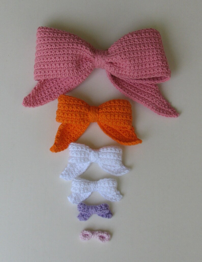 May include: A set of six crocheted bows in various sizes and colors, including pink, orange, white, purple, and light pink. The bows are arranged in order of size, with the largest bow at the top and the smallest bow at the bottom.