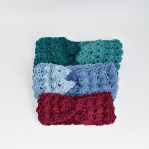 May include: Three crocheted headbands in different colors: teal, light blue, and burgundy. The headbands are stacked on top of each other.