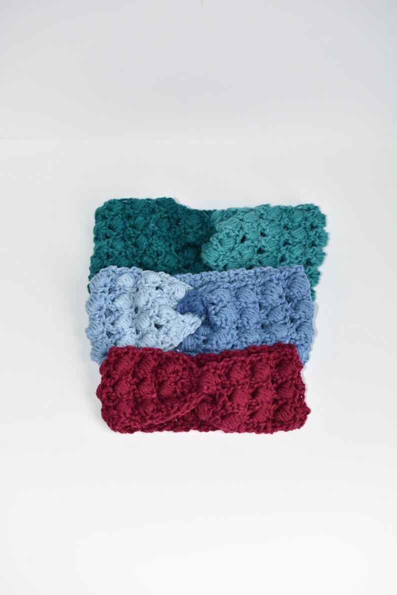 May include: Three crocheted headbands in different colors: teal, light blue, and burgundy. The headbands are stacked on top of each other.