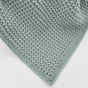 May include: A close-up of a crocheted blanket in a soft, muted green color. The blanket has a textured, raised stitch pattern, creating a dense and cozy appearance. The corner of the blanket is visible, showing the neat edge finishing.