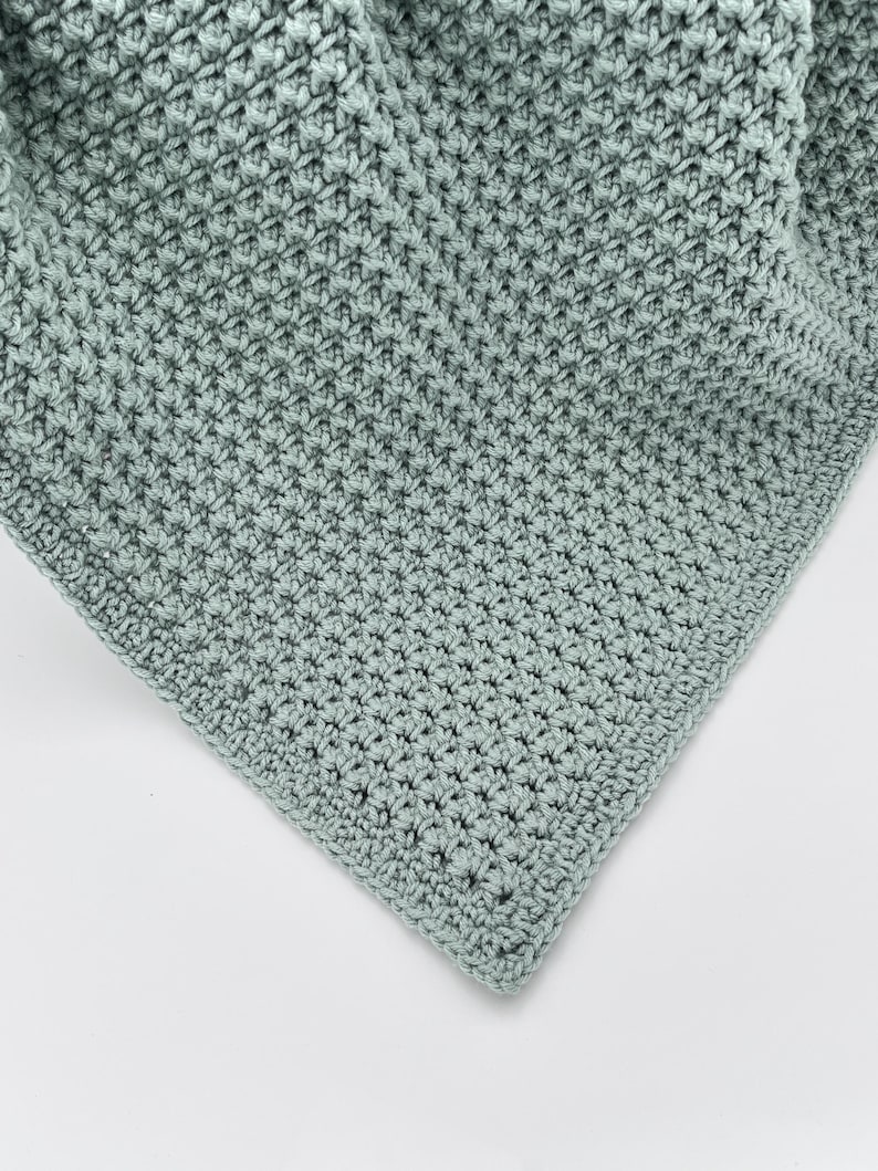 May include: A close-up of a crocheted blanket in a soft, muted green color. The blanket has a textured, raised stitch pattern, creating a dense and cozy appearance. The corner of the blanket is visible, showing the neat edge finishing.