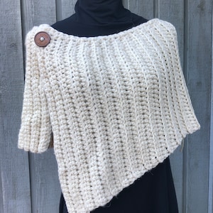 Pode incluir: Um poncho de crochê de cor creme com um botão de madeira no ombro.
