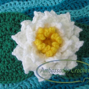 Puede incluir: Un nenúfar blanco de ganchillo con un centro amarillo, descansando sobre una hoja de nenúfar verde de ganchillo. La hoja de nenúfar está sobre un fondo de ganchillo a rayas azul y verde. El texto "Ambassador Crochet" es visible en la esquina inferior derecha de la imagen.