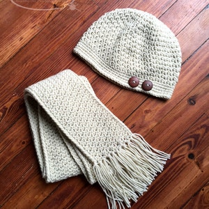 May include: A crocheted hat and scarf set in a cream color with brown buttons on the hat. The set is laid out on a wooden surface.