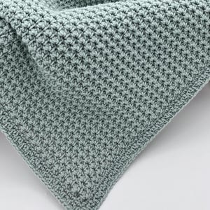May include: A close-up shot of a handmade, light teal crochet blanket. The blanket features a textured stitch pattern, creating a dense, warm appearance. The corner of the blanket is visible, showcasing the detailed craftsmanship and the soft, inviting texture of the yarn.