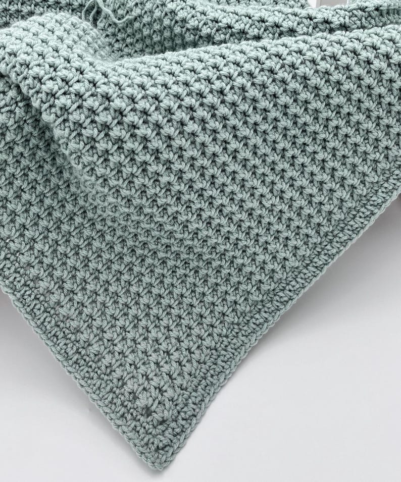 May include: A close-up shot of a handmade, light teal crochet blanket. The blanket features a textured stitch pattern, creating a dense, warm appearance. The corner of the blanket is visible, showcasing the detailed craftsmanship and the soft, inviting texture of the yarn.