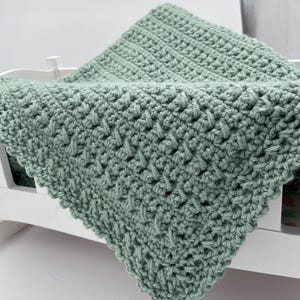 May include: A soft, sage green crocheted baby blanket with a delicate stitch pattern. The blanket is folded and draped over a white crib.
