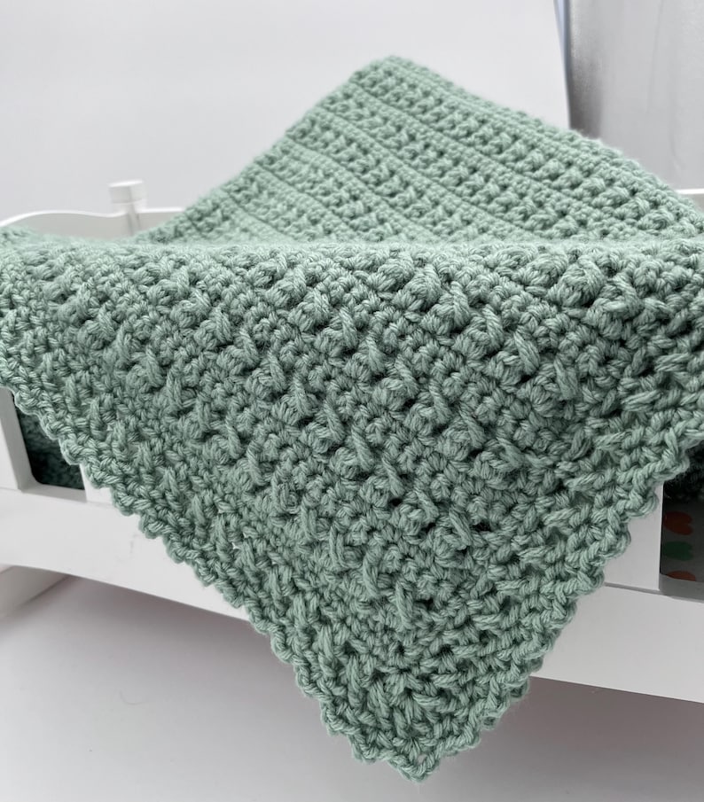 May include: A soft, sage green crocheted baby blanket with a delicate stitch pattern. The blanket is folded and draped over a white crib.