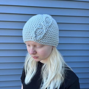 Op de afbeelding: Een lichtgrijze gehaakte muts met een kabelgebreid ontwerp aan de voorkant. De muts is gemaakt van zacht, getextureerd garen en is geschikt voor koud weer. Een stijlvol accessoire voor elke outfit.