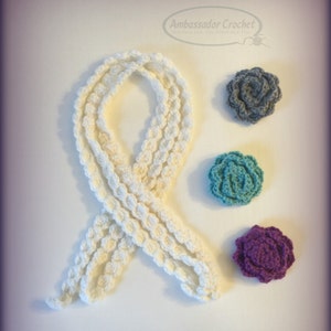 Könnte beinhalten: Ein weißes Häkelband mit drei gehäkelten Blumen in verschiedenen Farben: Grau, Türkis und Lila. Ambassador Crochet - Hooking Our World Together.
