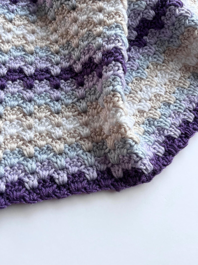 May include: A crocheted baby blanket in shades of purple, blue, and beige. The blanket has a textured stitch pattern and a scalloped edge.