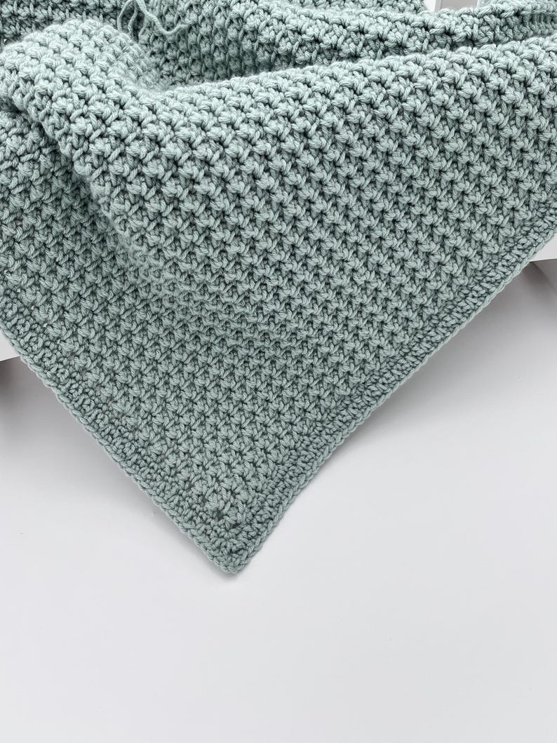 May include: A close-up of a handmade, light teal crochet blanket. The blanket features a textured stitch pattern, with a dense, woven appearance. The corner of the blanket is visible, showcasing the detailed craftsmanship and soft color.