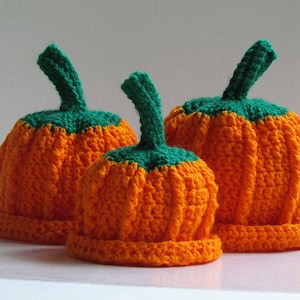 Puede incluir: Tres gorros de punto de calabaza de diferentes tamaños. Los gorros son de color naranja con tallos verdes.