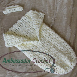 May include: A white crocheted baby sleeping bag with a matching headband. The sleeping bag has a scalloped edge and is made with a simple stitch pattern. The text "Ambassador Crochet, Reaching Out, One Stitch At a Time" is visible in the image.
