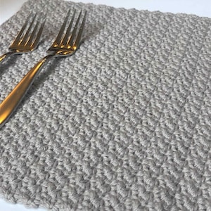 May include: A gray crocheted placemat with a subtle textured pattern. Two silver forks are placed on top of the placemat.