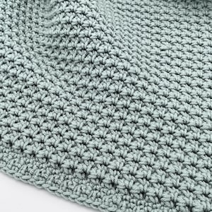 May include: Close-up of a crocheted blanket in a soft, muted green color. The blanket has a textured surface created by the tight stitches. The edges are neatly finished, and the fabric is folded, suggesting a cozy, handmade item.