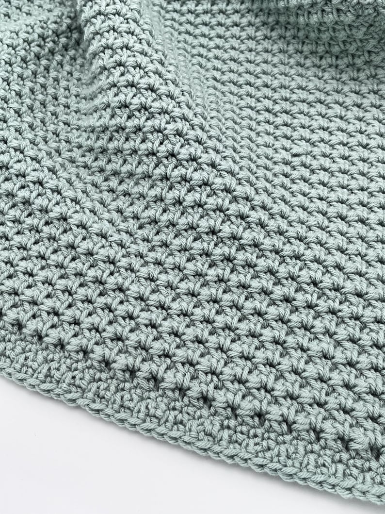 May include: Close-up of a crocheted blanket in a soft, muted green color. The blanket has a textured surface created by the tight stitches. The edges are neatly finished, and the fabric is folded, suggesting a cozy, handmade item.