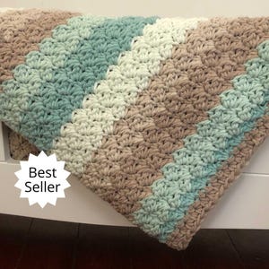 May include: A crocheted baby blanket with a chevron pattern in shades of beige, white, and teal. The blanket is draped over a white crib and has a "Best Seller" tag attached.